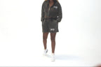 A model wearing a dark gray zip-up hoodie and matching shorts set with white sneakers, standing against a plain white studio background.
