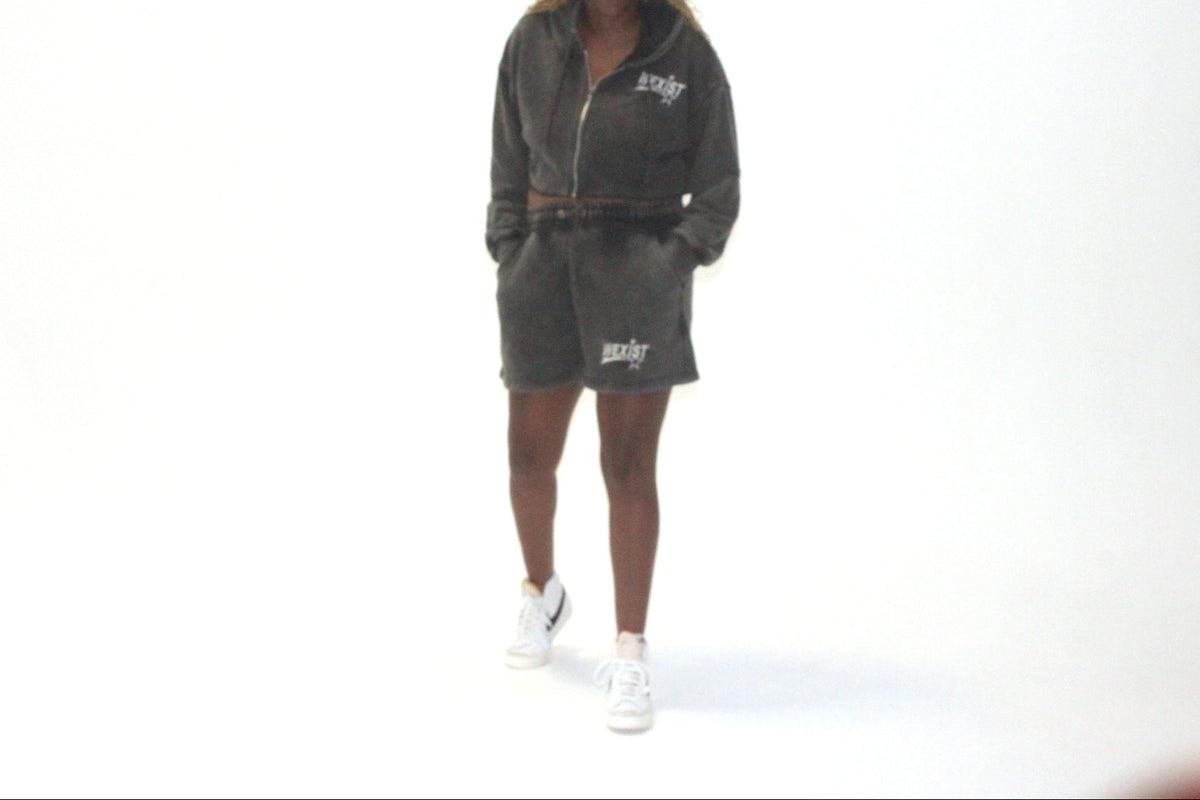 A model wearing a dark gray zip-up hoodie and matching shorts set with white sneakers, standing against a plain white studio background.