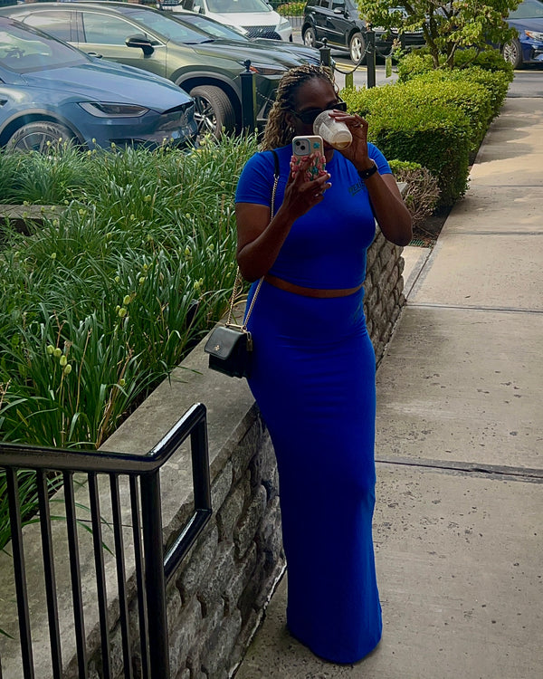 A tall woem woman wearing a bright royal blue two-piece dress stands on a sidewalk, holding a phone and a cup. The dress has short sleeves, a fitted bodice with a thin brown belt at the waist, and a floor-length skirt.