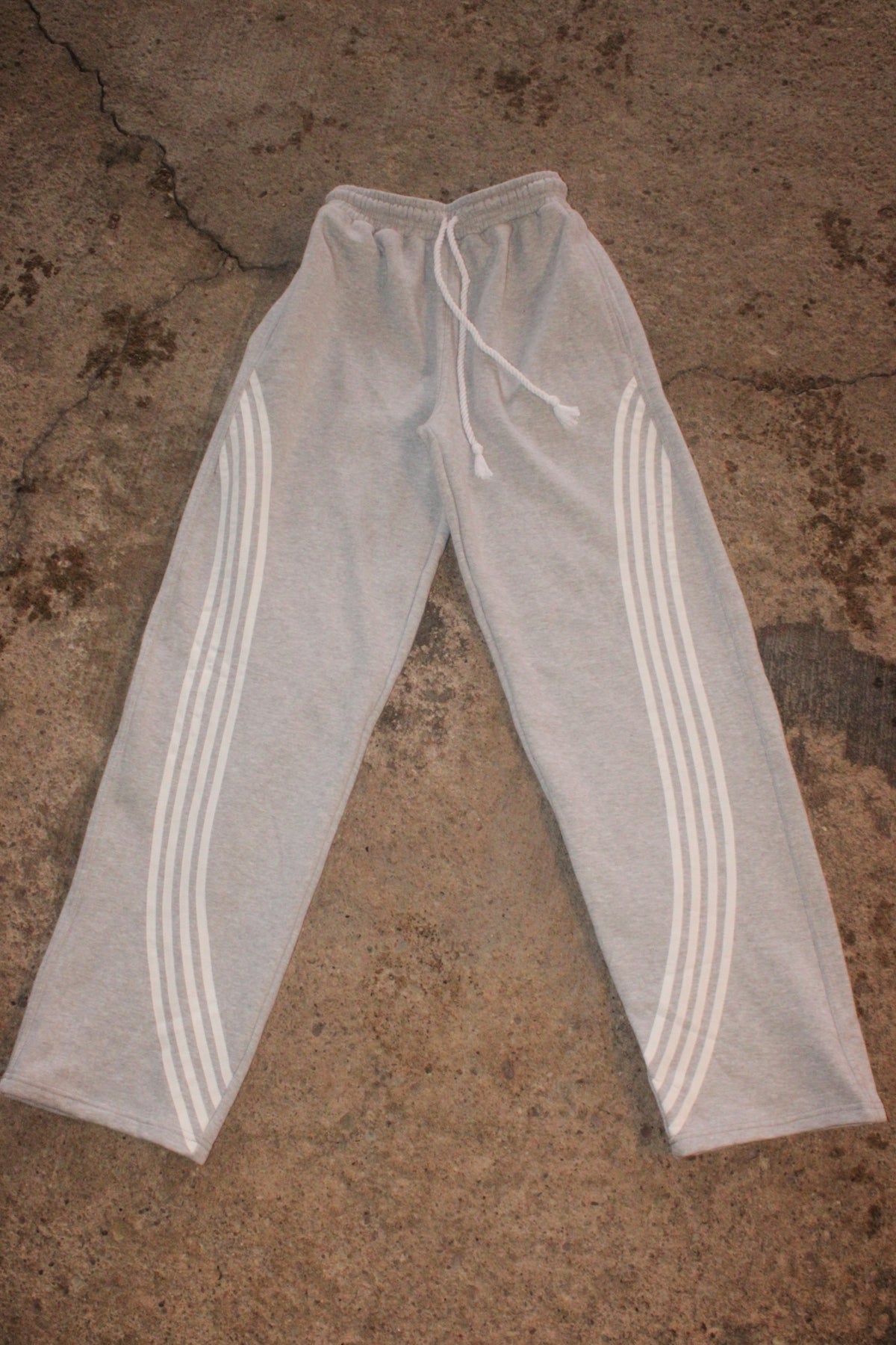 Light gray heather sweatpants with three white stripes down each leg and a white drawstring waist, laid flat on a textured concrete surface. WEXIST gray tall girl varsity cotton fleece joggers